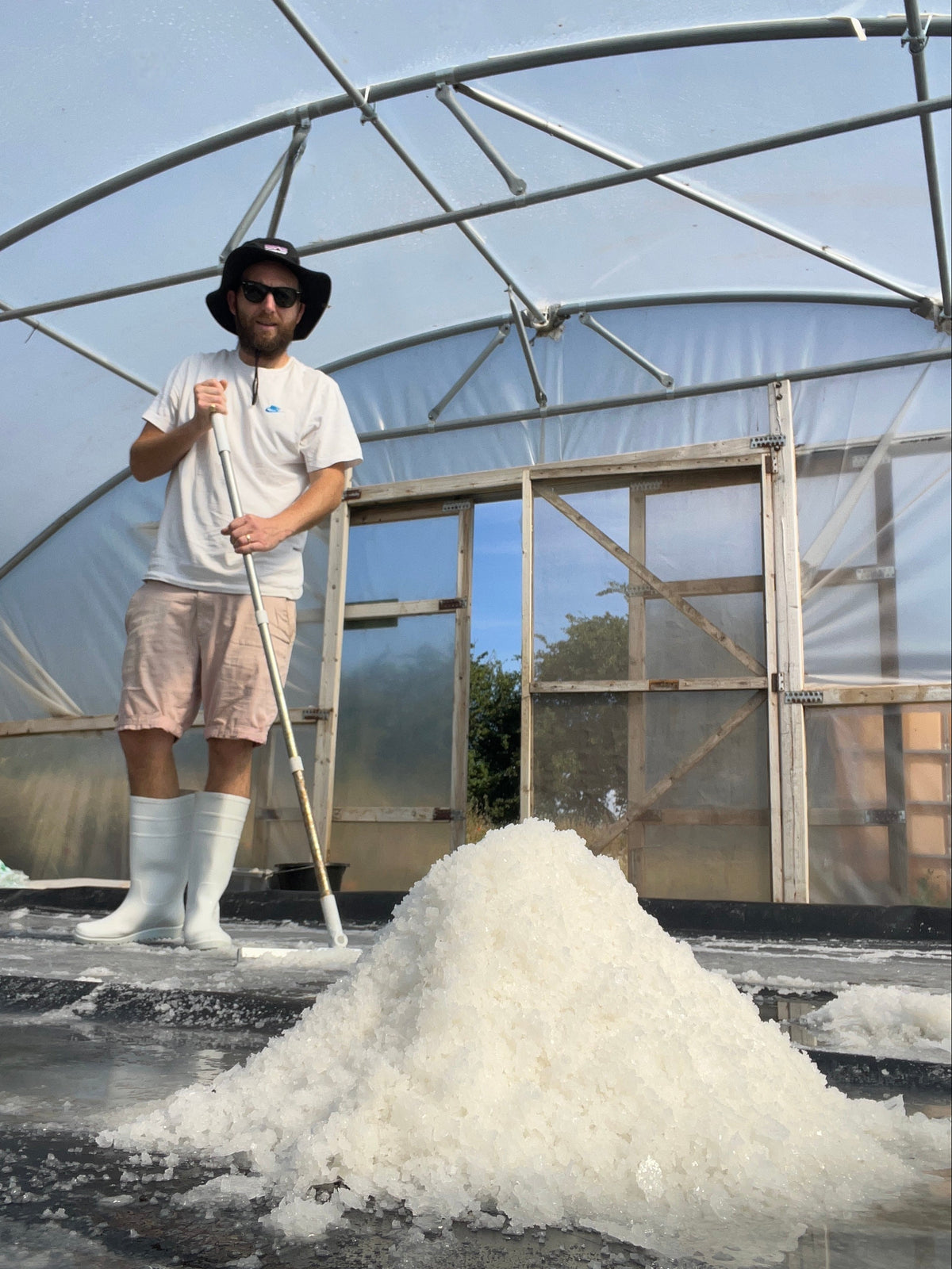 A man wearing a white t-shirt, light pink shorts, white wellington boots and a black bucket hat stands inside a polytunnel holding a long-handled tool next to a large pile of white Suffolk Sea Salt.