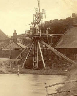 Sepia-toned vintage photograph showing a large wooden wind pump wheel beside a river, with buildings and trees in the background.