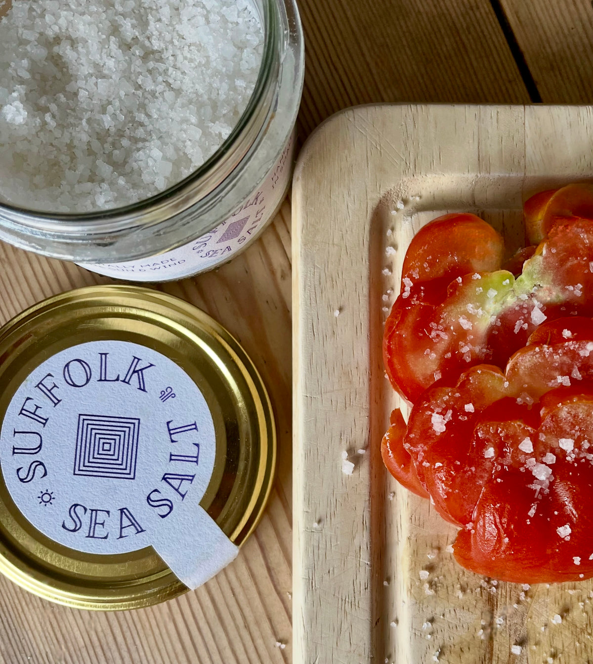 Open jar of Suffolk Sea Salt beside its gold lid and sliced tomatoes sprinkled with sea salt on a wooden board.