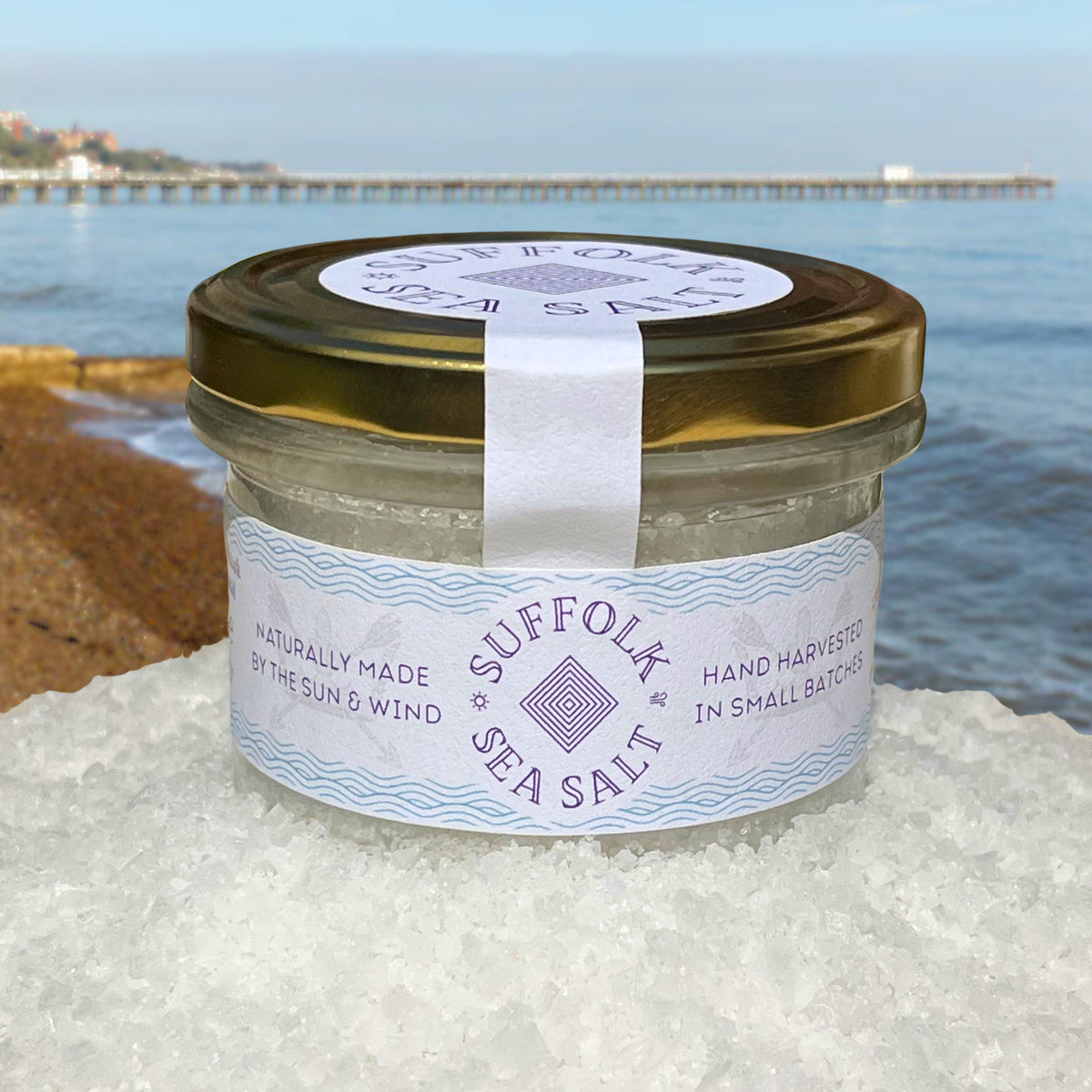 Suffolk Sea Salt jar with a gold lid, sitting on sea salt with the sea and Felixstowe pier in the background.